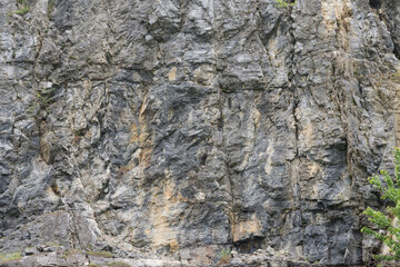 Weathered rock face texture in old stone pit with parts of green. Aged stone wall surface background pattern with cracks and scratches