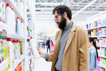 Young brunette man with glasses with a beard in a supermarket chooses a cleaning product for the...