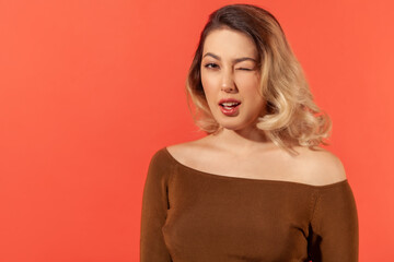 Fototapeta premium Portrait of flirting playful young woman with blonde hair in brown blouse, winking and smiles. Share love and friendship feelings, looking at camera. Indoor studio shot isolated on red background
