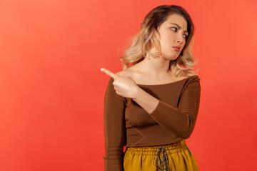 Get out! Irritated upset blonde woman in brown blouse pointing away, quarreling and scolding, showing exit with angry grimace, feeling betrayed. Indoor studio shot isolated on red background