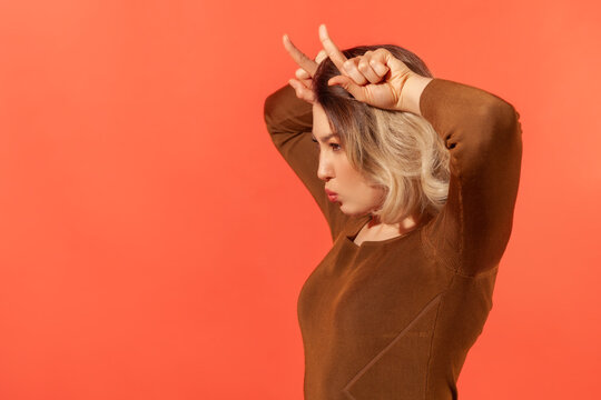 Side view portrait of angry menacing blonde girl in brown blouse showing bull horns on head, threatening to attack, danger and aggression concept. Indoor studio shot isolated on red background