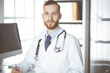 Friendly red-bearded doctor sitting and writing at clipboard in clinic. Perfect medical service in hospital. Medicine concept