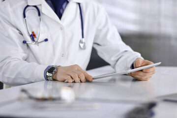 Unknown male doctor sitting and working with tablet computer in clinic at his working place, close-up. Young physician at work. Perfect medical service, medicine concept