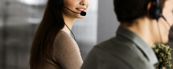 Young cheerful girl in headsets is looking at camera, while sitting at the desk with a male...