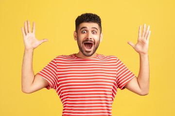 Shocked surprised bearded man in red striped t-shirt looking at camera with big eyes and widely opened mouth, amazed and excited. Indoor studio shot isolated on yellow background