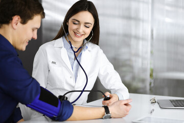 Young woman-doctor is checking her patient's blood pressure, while they are sitting together at the desk in a cabinet. Physician at work in a hospital. Medicine and healthcare concepts