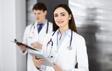 Smiling intelligent woman-doctor is holding a tablet computer in her hands, while she is standing in a clinic with a colleague at the background. Portrait of physician at work. Perfect medical service