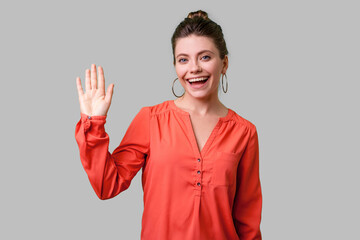 Hello! Portrait of charming woman with bun hairstyle, big earrings and in red blouse standing, waving hand and saying Hi to camera with toothy smile. indoor studio shot isolated on gray background