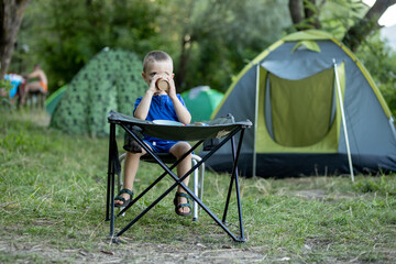 Little boy eating breakfast outdoor at camping site in nature sunlight with a tent at the background.