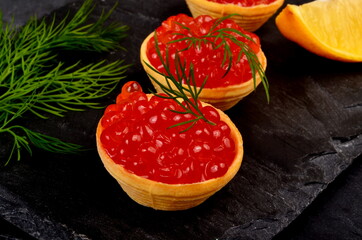 Tartlets with red salmon caviar on black stone plate, decorated with dill and a slice of lemon, close - up. Seafood appetizer