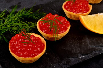 Tartlets with red salmon caviar on black stone plate, decorated with dill and a slice of lemon, close - up. Seafood appetizer