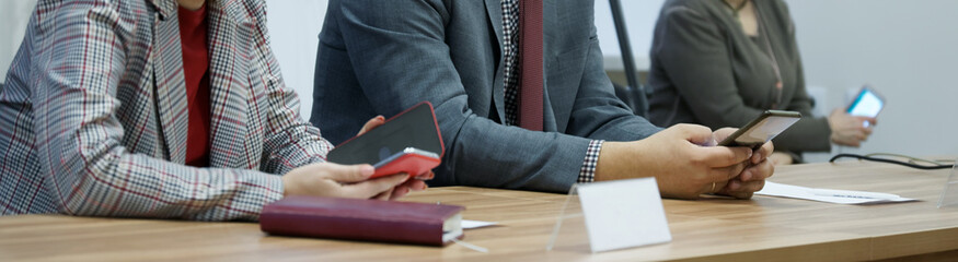 Sitting at a table during a meeting or negotiation, men and women use smartphones. Modern...