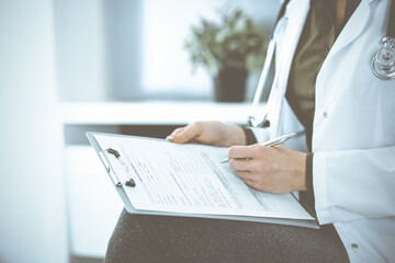 Unknown woman-doctor writing something at clipboard while sitting at the chair, close-up. Therapist wearing green blouse at work is filling up medication history record. Medicine concept