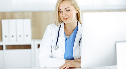 Woman-doctor at work while sitting at the desk in hospital or clinic. Blonde cheerful physician using desktop computer
