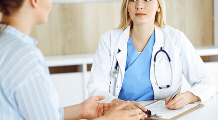 Woman-doctor and patient sitting and talking in hospital or clinic. Blonde therapist is cheerfully...