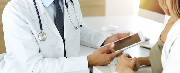 Doctor and patient discussing medical exam results while sitting at the desk in sunny clinic, close-up. Male physician using tablet computer for filling up medication history record.