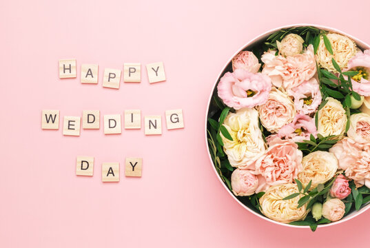 Happy wedding day lettering on the wooden squares with letters on the pink background and big round box with red flowers and roses.