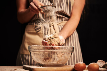 Female baker hands making dough for bread with an apron. Natural homemade ingredients. Dark background, brown color grading.