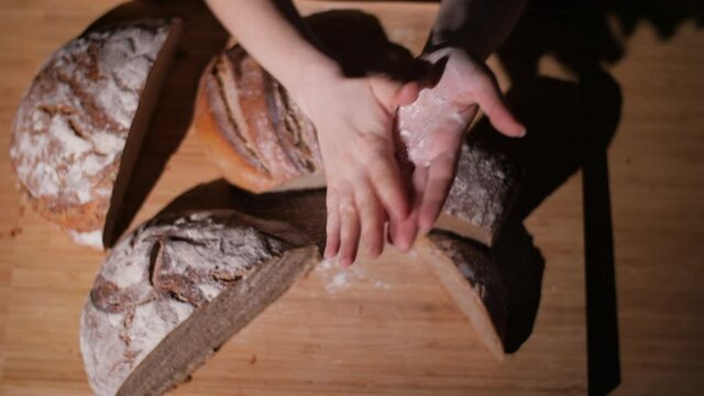 Top-down view on the hands of a home baker dusting off flour. Sliced rustic whole-wheat bread loaves on a wooden cutting board in the background. 4K.