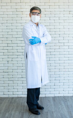 Portrait of full length Asian elder doctor or researcher wear lab coat, clear eyeglasses and face mask standing and arm crossed with white brick background.