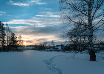 sunrise over the village. pink-blue sky with white floating clouds, white snow with a trampled path, silhouettes of trees and wooden houses