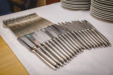 many clean steel knives lie in a row line on a tablecloth in a restaurant with plates on the background