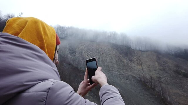 Woman traveler in yellow hoodie taking photos of evaporation at the mountain cliff using smartphone. Female tourist photographing beautiful nature at cold weather.