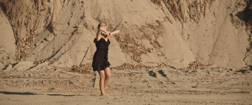 Charming cute slim brunette girl in black short dress makes beautiful dance moves in the form of latino on the sand on background of sand hill on sunny day