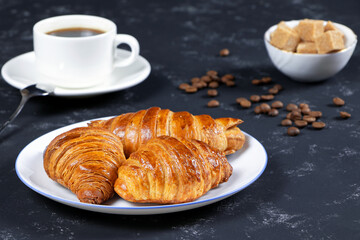 A cup of hot coffee and croissants on a black background. Breakfast with coffee and fresh pastry. Coffee beans on a table. Fresh cake and pastry