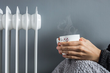 Hands holding a cup of hot drink, in front of the heater with a sheepskin blanket. Relaxed woman in her home