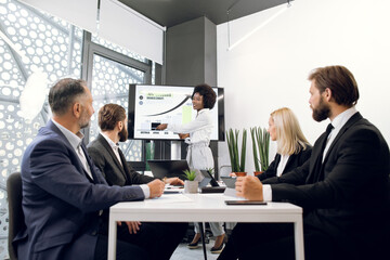 Beautiful confident African American black-skinned lady, showing her financial ad sales report on big digital screen for her focused high-skilled multiracial business colleagues
