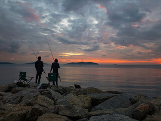 Two fisherman with chairs and rods standing on rocks during sunset and trying to catch some fishes. A cat lies beside them and waiting for food. Cloudy and dramatic sky background with some islands