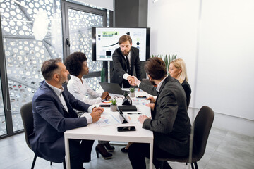 Handsome confident bearded young businessman boss shaking hand with his young male colleague, preparing for the meeting and negotiations with multiethnic colleagues