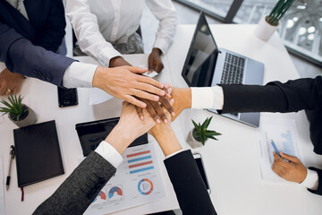 Close up top view of diverse multiethnical business people putting their hands together, showing unity. Diverse team joining stacking their hands. Table with laptop, tablets, papers