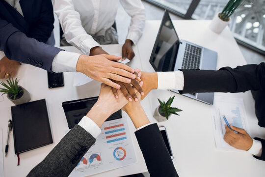 Close up top view of diverse multiethnical business people putting their hands together, showing unity. Diverse team joining stacking their hands. Table with laptop, tablets, papers