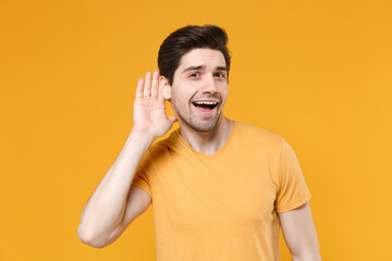 Young curious unshaved caucasian handsome man years old wearing casual basic blank print design t-shirt try to hear you overhear listening intently isolated on yellow color background studio portrait.
