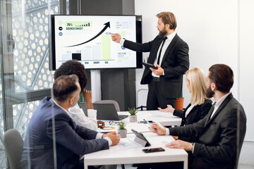Front view of young confident bearded businessman, showing statistic datas on big plasma screen for his focused high-skilled multiethnical colleagues during meeting in office room