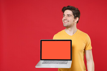 Young smiling unshaved man 20s in basic yellow t-shirt using hold laptop pc computer with blank screen workspace copy space mock up looking aside isolated on red color background studio portrait.