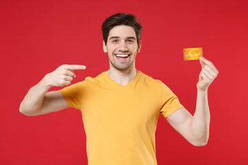 Young smiling unshaved successful gladden happy man 20s wearing casual basic yellow t-shirt holding in hand pointing index finger on credit bank card isolated on red color background studio portrait.