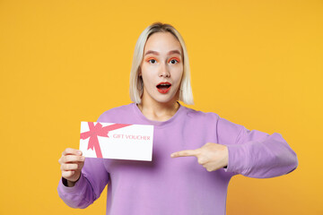 Fototapeta na wymiar Shocked young blonde caucasian woman 20s with bob haircut bright makeup wearing casual basic purple shirt hold pointing finger on gift voucher isolated on yellow color background studio portrait.