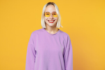 Smiling fun young blonde fascinating caucasian woman 20s bob haircut bright makeup wearing basic casual purple shirt and glasses looking camera isolated on yellow color background studio portrait.