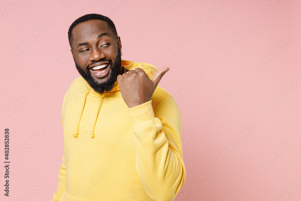 Wall mural Side view of funny young african american man 20s in casual basic yellow streetwear hoodie standing pointing thumb aside on mock up copy space isolated on pastel pink color background studio portrait.