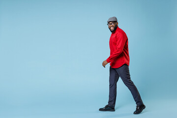 Full length side view of smiling cheerful young bearded african american man in casual red shirt cap eyeglasses standing walking going looking aside isolated on pastel blue background studio portrait. - Powered by Adobe
