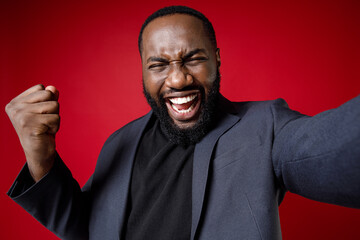 Close up of joyful young african american business man 20s in classic jacket suit standing doing selfie shot on mobile phone doing winner gesture isolated on red color background studio portrait.