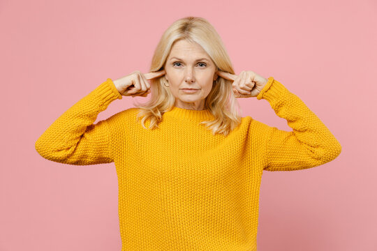 Concerned Elderly Gray-haired Blonde Woman Lady 40s 50s Years Old In Yellow Casual Sweater Standing Covering Ears With Fingers Looking Camera Isolated On Pastel Pink Color Background Studio Portrait.