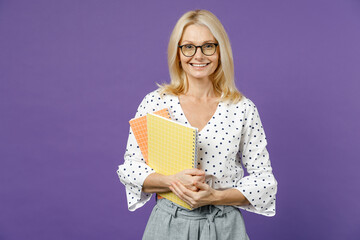 Smiling gray-haired blonde teacher woman lady 40s 50s years old wearing white dotted blouse eyeglasses standing hold notepads looking camera isolated on bright violet color background studio portrait. - obrazy, fototapety, plakaty