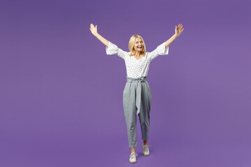 Full length of excited funny elderly gray-haired blonde woman lady 40s 50s in white dotted blouse standing rising spreading hands looking camera isolated on violet color background studio portrait.