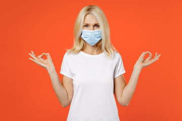 Elderly gray-haired blonde woman lady 40s 50s in t-shirt face mask safe from coronavirus virus covid-19 hold hands in yoga gesture relaxing meditating try to calm down isolated on orange background.