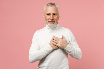 Smiling elderly gray-haired mustache bearded man wearing casual basic white turtleneck standing holding hands folded on heart looking camera isolated on pastel pink wall background studio portrait.