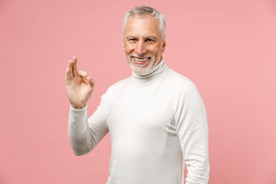 Smiling cheerful elderly gray-haired mustache bearded man wearing casual basic white turtleneck standing showing OK gesture looking camera isolated on pastel pink color background studio portrait.